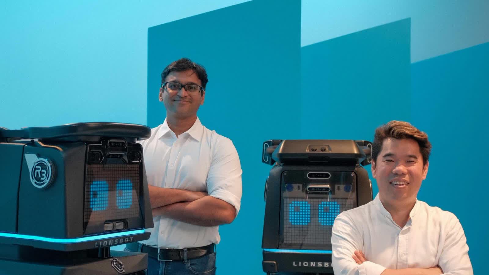 Two members of the LionsBot team standing with their arms folded between two R3 cleaning robots against a blue studio backdrop