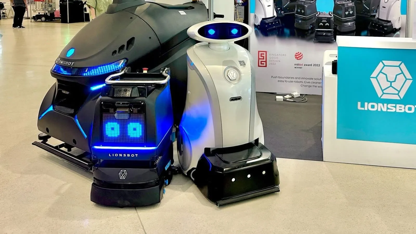 Three LionsBot cleaning robots of different sizes grouped together on the stand at NRP 6, among them a white LeoBot with its digital face lit, in front of a LionsBot banner carrying its 2022 design award marks