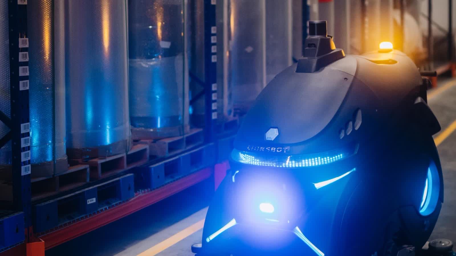 The R12 Rex CS working a warehouse aisle at night, its blue light bars and amber roof beacon lit, beside racking stacked with pallets