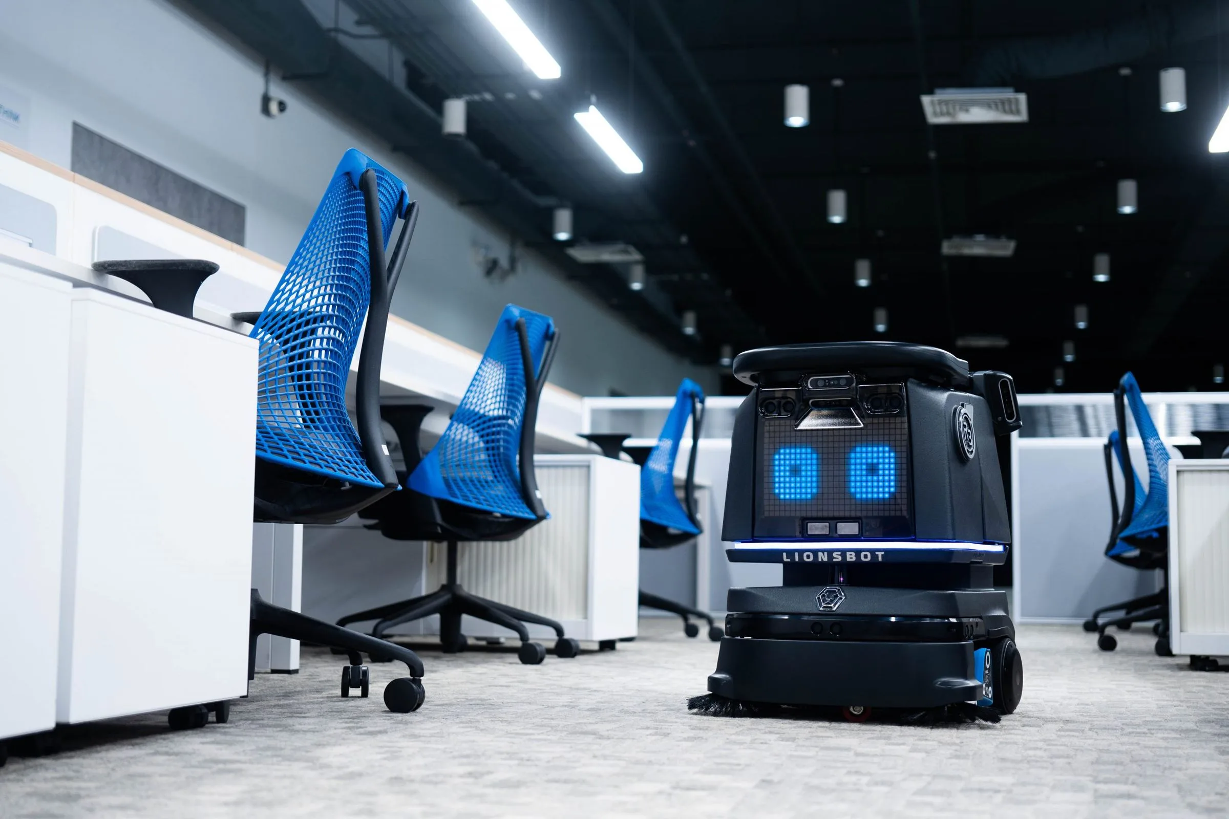 A LionsBot R3 Vac autonomous vacuum cleaning the carpet between desks in an open-plan office