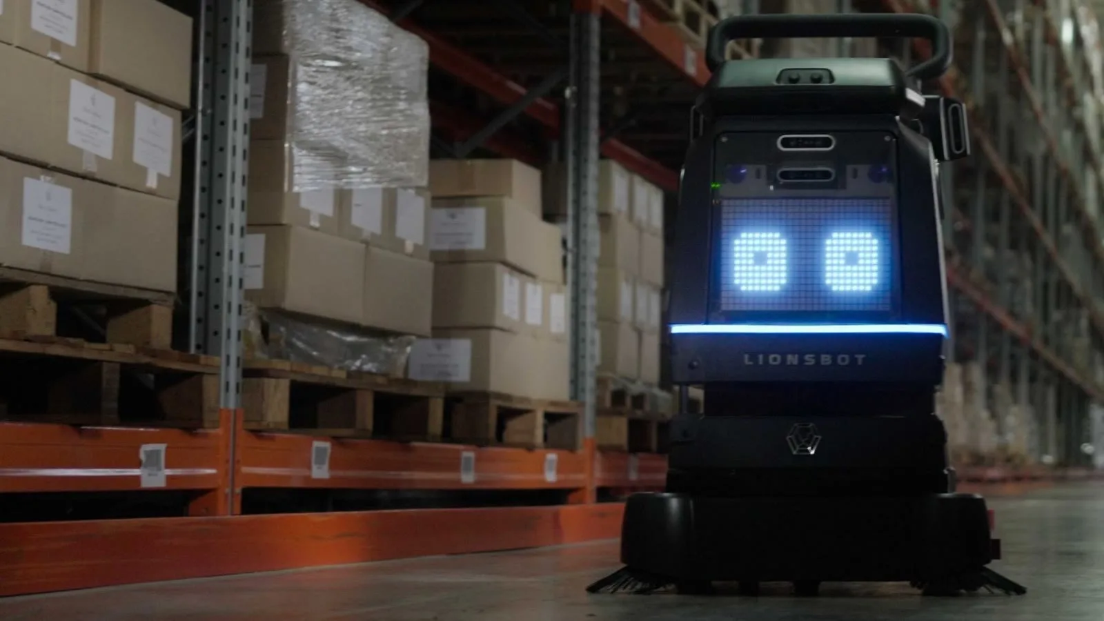 A LionsBot R3 Scrub Pro autonomous floor scrubber in a warehouse aisle, its display lit, stacked pallet racking either side