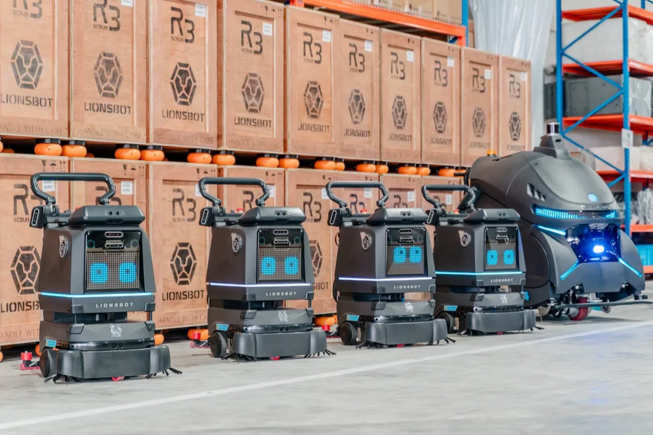 Four R3 Scrub Pro robots lined up beside a larger Rex robot, in front of stacked wooden LionsBot shipping crates in the factory warehouse