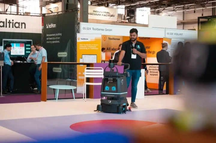 A LionsBot cleaning robot working along an aisle of the show floor while a team member walks beside it