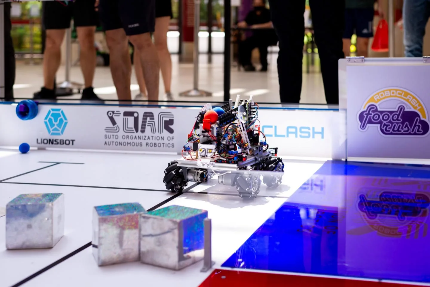 A competition robot mid-run on the RoboClash playfield, pushing a patterned cube, with SOAR and LionsBot banners behind it