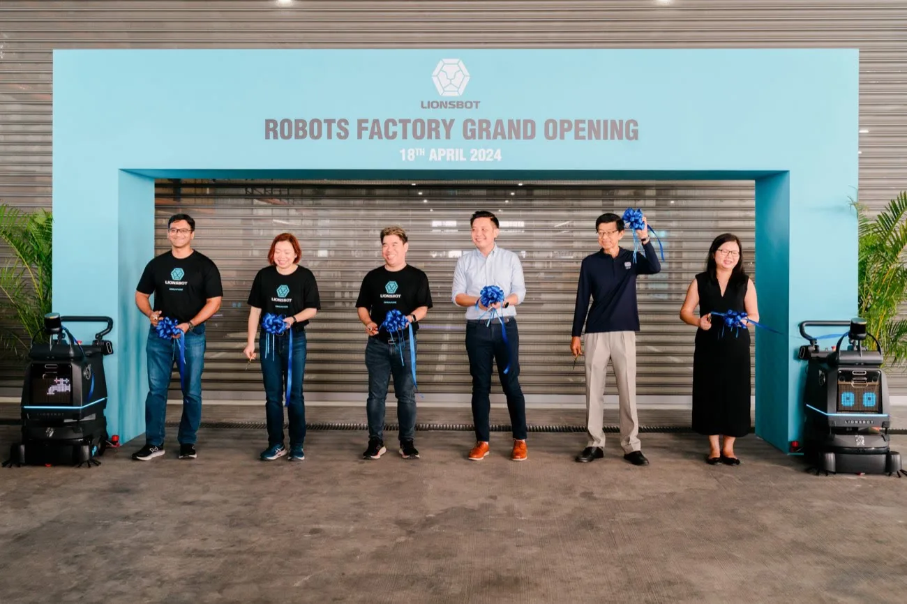 Six LionsBot and guest representatives holding a blue ribbon in front of a backdrop reading Robots Factory Grand Opening, 18th April 2024, with a LionsBot robot standing at each end