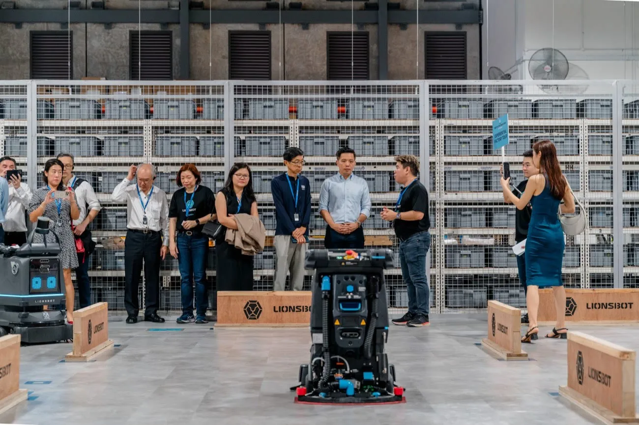 Guests standing in the factory warehouse watching a LionsBot scrubber robot being demonstrated on the floor in front of shelves of parts bins