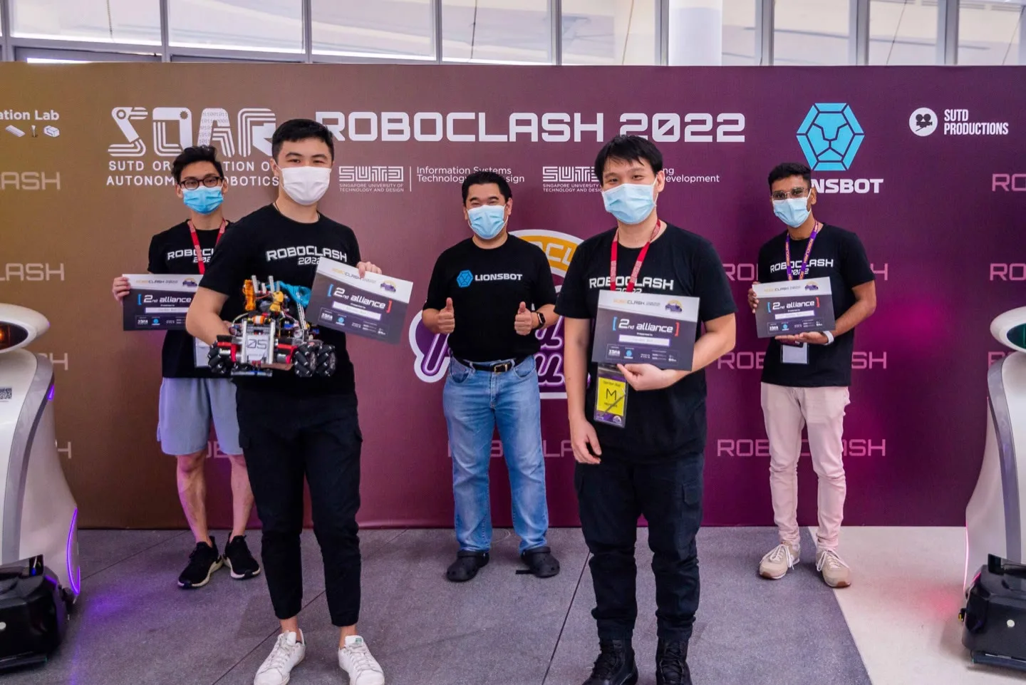 Five RoboClash 2022 participants holding prize cheques in front of a RoboClash 2022 backdrop carrying the SOAR and LionsBot logos