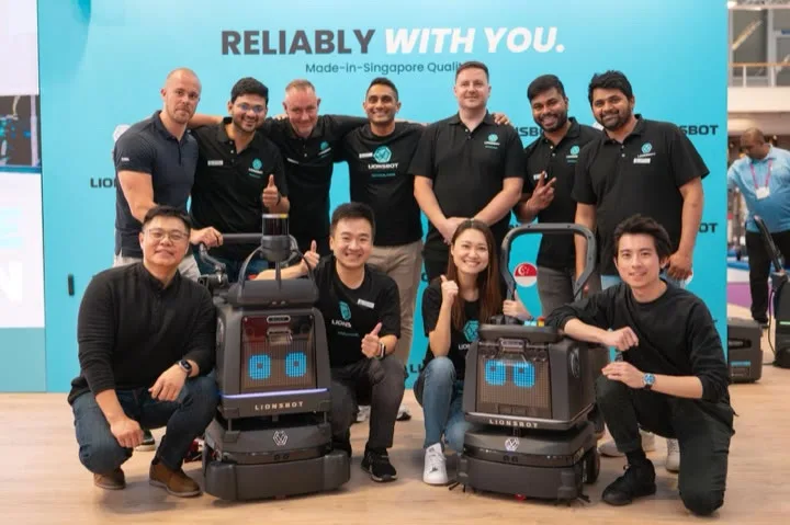 Eleven members of the LionsBot team posed with two cleaning robots in front of a stand banner reading "Reliably with you. Made-in-Singapore Quality"