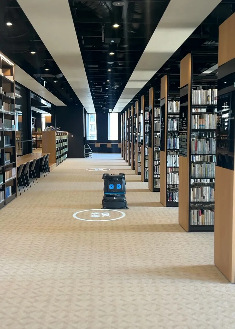 A LionsBot R3 Vac working down a library aisle between tall bookshelves, its guide markers projected on the carpet ahead