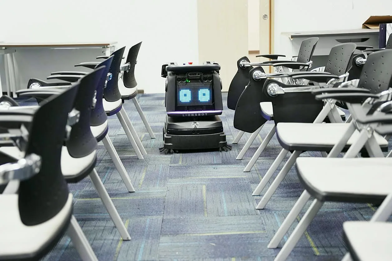 A LionsBot R3 Vac vacuuming a carpeted lecture room between rows of fixed seating
