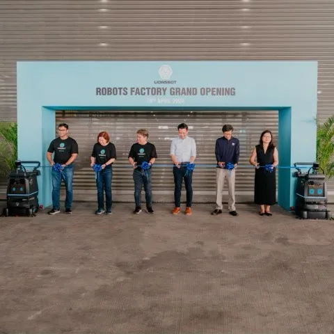 Six people cutting a ribbon at the LionsBot robots factory grand opening, with a robot standing at each side of the entrance