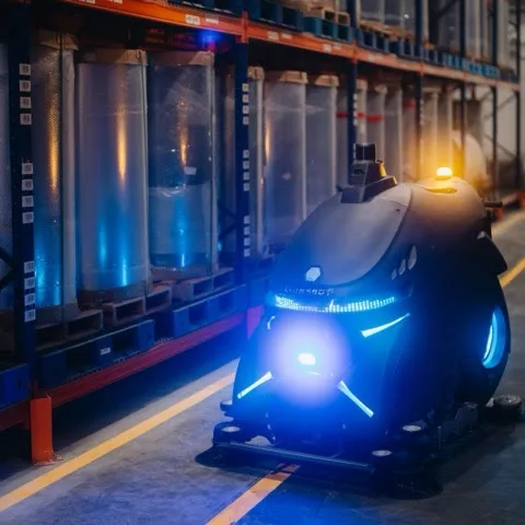 The R12 Rex CS cleaning a warehouse aisle, its blue light bars reflected on the floor