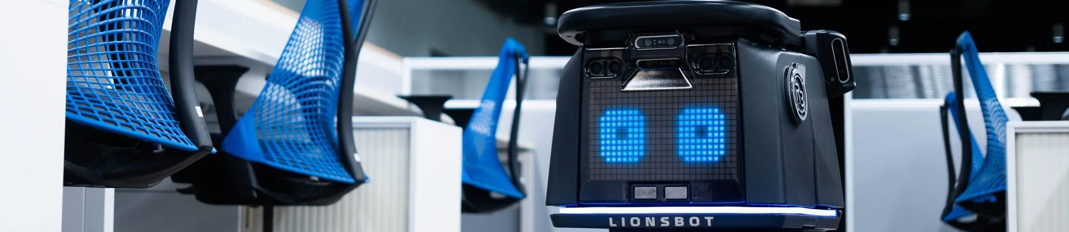 A LionsBot R3 Vac vacuuming the carpet beside a row of office desks and chairs
