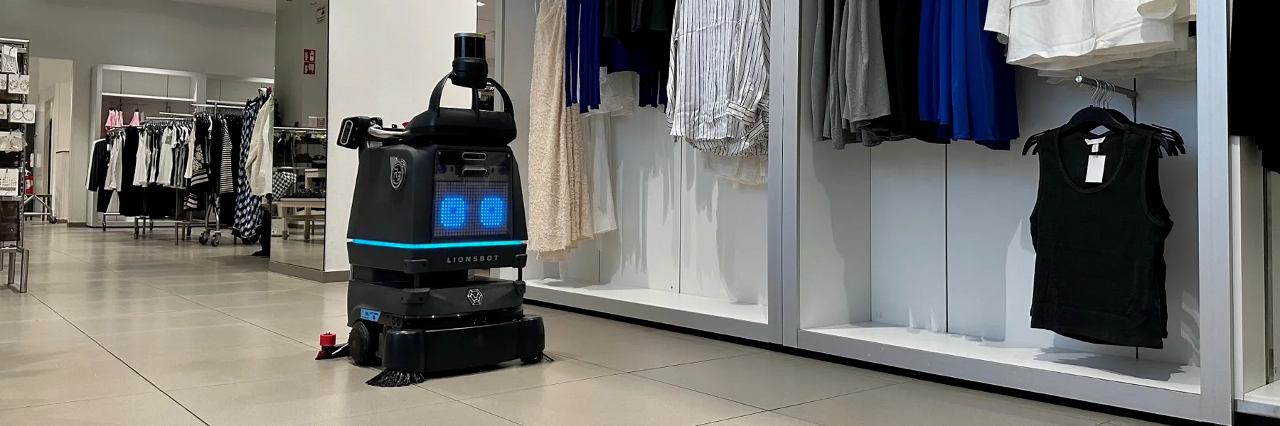 A LionsBot R3 Scrub Pro scrubbing the floor of a retail store aisle during opening hours, beside racks of clothing