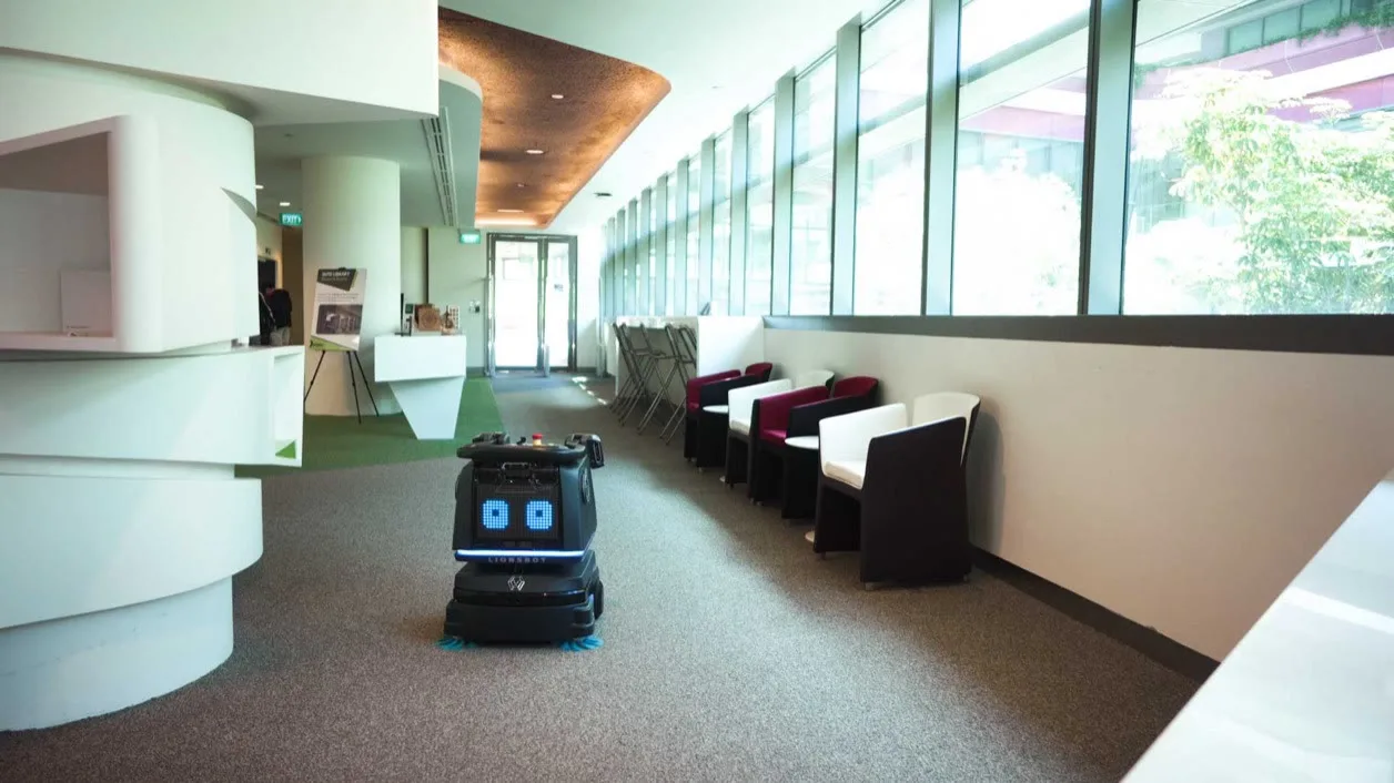 LionsBot R3 Vac vacuuming along a carpeted corridor lined with seating