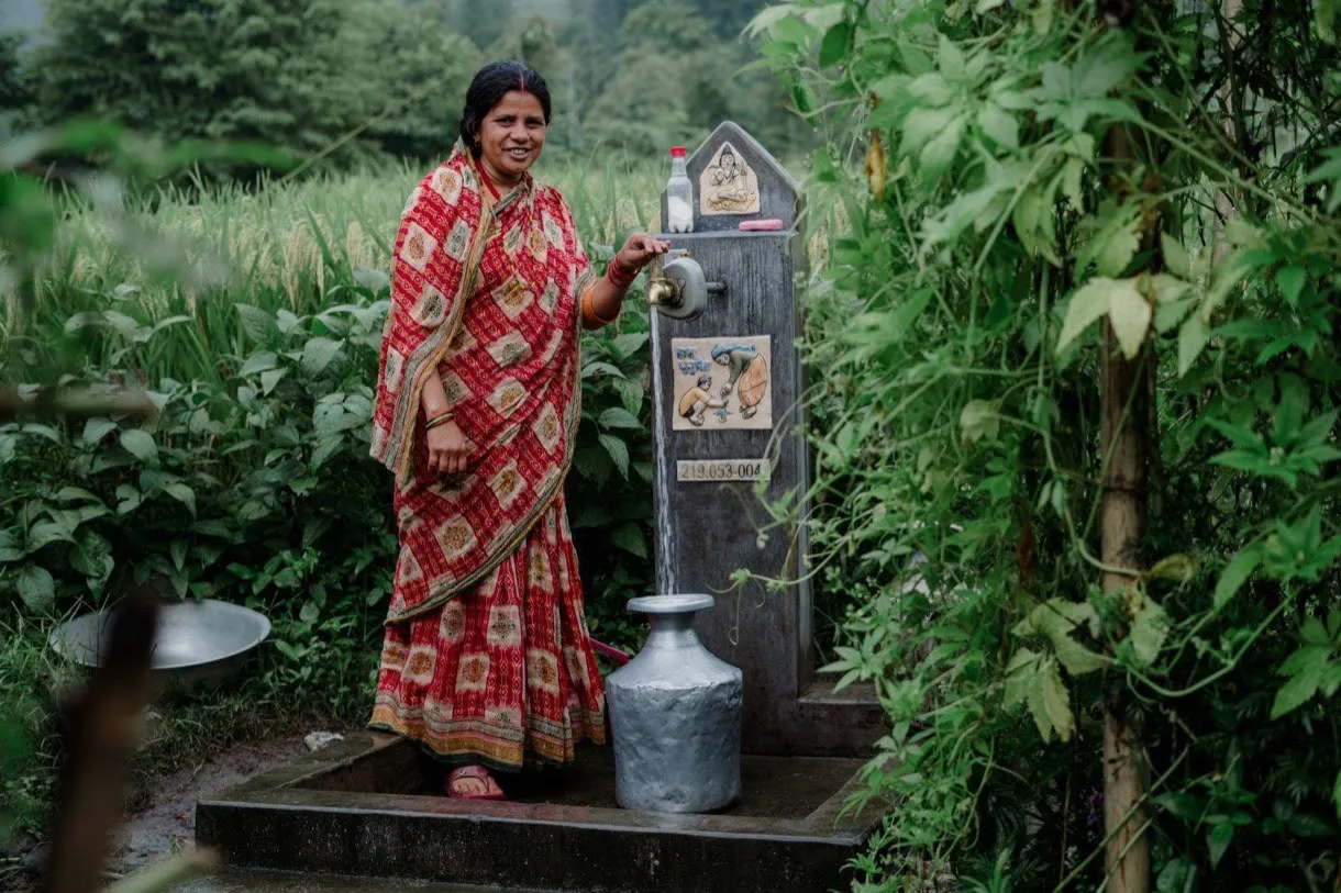 A woman in a red patterned sari standing at a village tap stand surrounded by green fields, filling a metal water pot