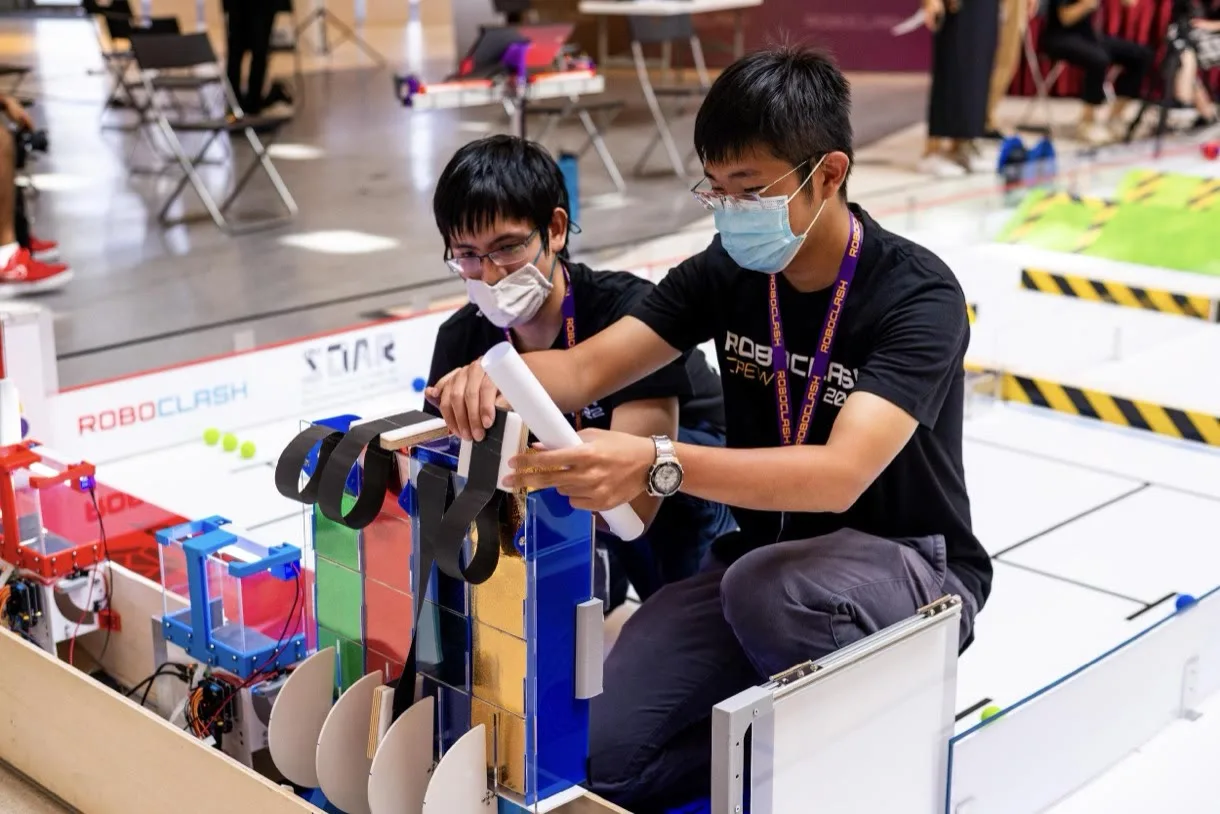Two RoboClash crew members in competition T-shirts assembling a student-built robot beside the contest arena