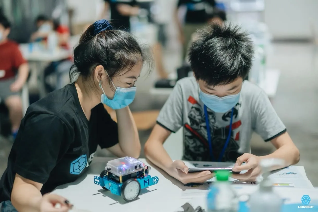 A LionsBot volunteer in a branded T-shirt helping a child programme a small wheeled robot at a workshop table