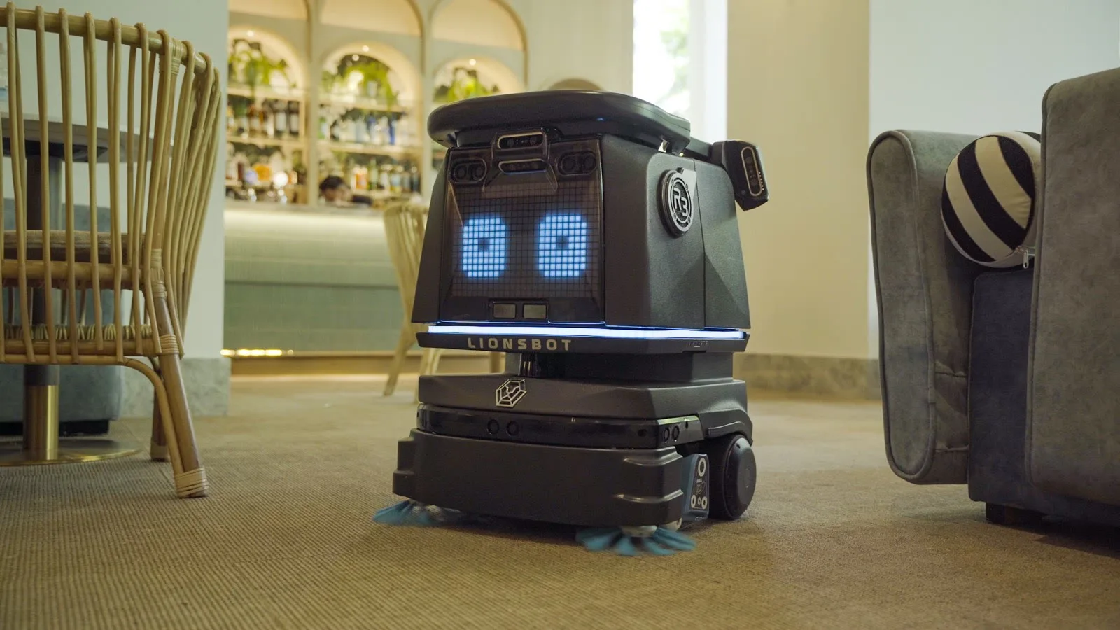 A LionsBot R3 Vac autonomous vacuum cleaning the carpet of a cafe, passing between a rattan chair and an armchair with the bar counter behind